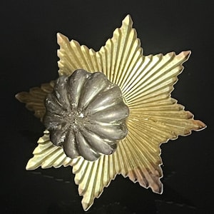 Könnte beinhalten: Nahaufnahme eines sternförmigen Dekorationsgegenstands. Das Objekt hat eine silberfarbene, abgerundete Mitte und eine goldfarbene, sternförmige Außenschicht mit geriffelten Details. Der Hintergrund ist schwarz.