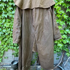 May include: A long, brown waxed cotton coat with a cape-like collar. The coat has a button closure and is designed to provide protection from the elements. The coat is hanging against a backdrop of green ivy and a brick wall.