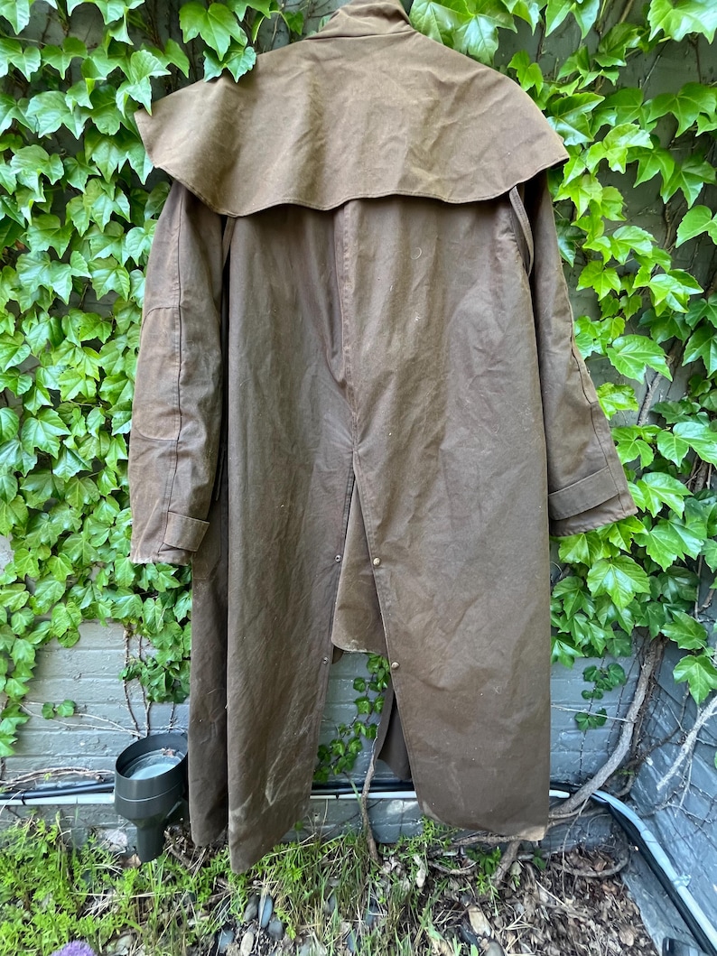 May include: A long, brown waxed cotton coat with a cape-like collar. The coat has a button closure and is designed to provide protection from the elements. The coat is hanging against a backdrop of green ivy and a brick wall.