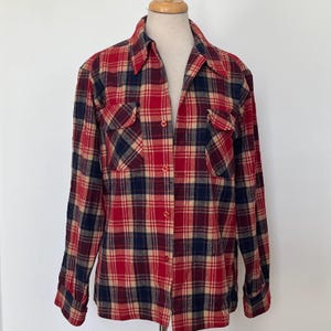 Peut inclure: Une chemise en flanelle à carreaux rouges, bleu marine et beige, avec deux poches de poitrine et un col. La chemise à manches longues est boutonnée sur le devant et présentée sur un mannequin.