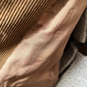 Op de afbeelding: Close-up van bruine corduroy stof met een geribbelde textuur. Grenzend aan de corduroy is een lichtroze satijnen voering, met een zichtbare naad. De afbeelding suggereert een kledingstuk, mogelijk een jas of jas.