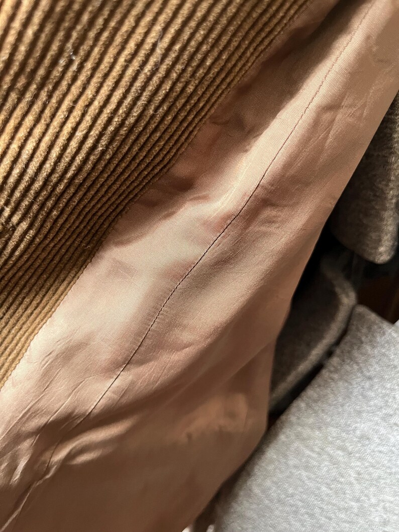 Op de afbeelding: Close-up van bruine corduroy stof met een geribbelde textuur. Grenzend aan de corduroy is een lichtroze satijnen voering, met een zichtbare naad. De afbeelding suggereert een kledingstuk, mogelijk een jas of jas.