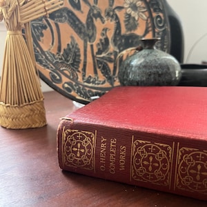 May include: A red hardcover book titled "O. Henry Complete Works" with gold lettering and decorative patterns. A straw figure and a decorative plate with animal figures are in the background.