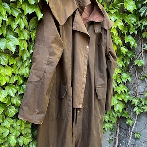 May include: A brown waxed cotton coat with a hood, hanging against a backdrop of green ivy. The coat has a long length, button closures, and pockets. The color is a deep, earthy brown, suggesting durability and outdoor wear.