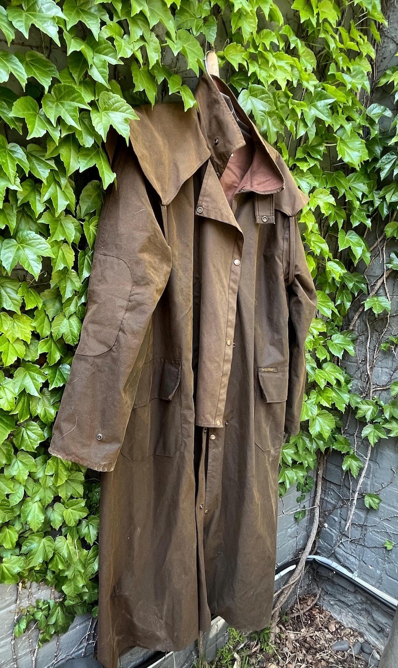 May include: A brown waxed cotton coat with a hood, hanging against a backdrop of green ivy. The coat has a long length, button closures, and pockets. The color is a deep, earthy brown, suggesting durability and outdoor wear.