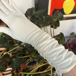 Peut inclure: Un gant long en cuir blanc avec un design froncé sur l'avant-bras. Le gant est d'un blanc uni et brillant et semble être fait d'un matériau lisse. Le gant s'étend au-delà du poignet.