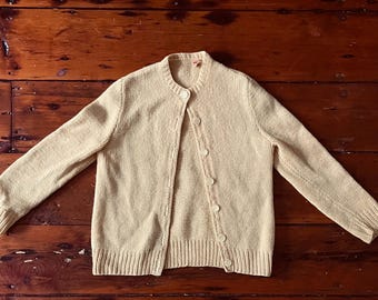 Pode incluir: Um cardigan de malha bege claro com fecho de botões. O cardigan tem uma textura de malha canelada e é feito de um material macio e confortável.