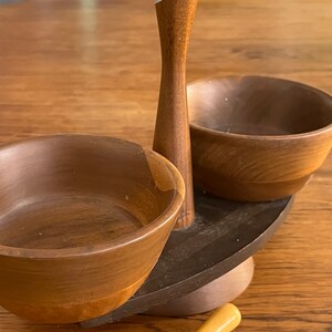 May include: A wooden serving set with two bowls and a central stand. The bowls are brown and the stand is dark brown with a light brown top. The set is on a wooden table.