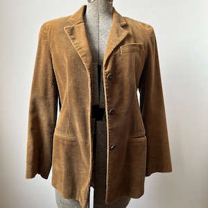 Op de afbeelding: Bruine corduroy blazer met een reverskraag, twee voorzakken en twee knopen. De jas wordt getoond op een vintage paspop. De corduroy stof heeft verticale ribbels.