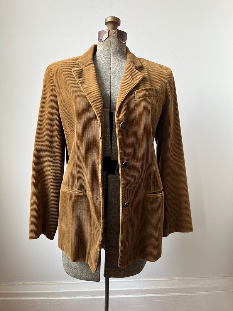 Op de afbeelding: Bruine corduroy blazer met een reverskraag, twee voorzakken en twee knopen. De jas wordt getoond op een vintage paspop. De corduroy stof heeft verticale ribbels.