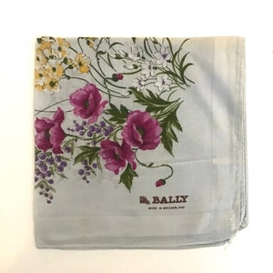Pu&ograve; includere: Un fazzoletto di seta blu chiaro con un motivo floreale con papaveri rosa, fiori bianchi e foglie verdi. Il fazzoletto ha un logo Bally marrone con il testo "MADE IN SWITZERLAND".
