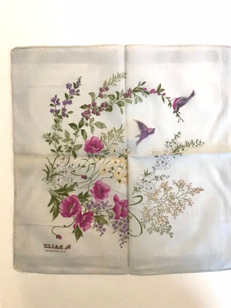 Pu&ograve; includere: Un foulard di seta blu chiaro con un motivo floreale che presenta papaveri rosa, foglie verdi e fiori bianchi. Due piccoli uccelli volano nell'angolo in alto a destra del foulard. Il testo "BAILLY" &egrave; stampato nell'angolo in basso a sinistra del foulard.