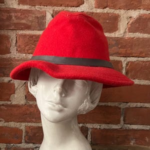 Puede incluir: Un sombrero fedora de fieltro rojo con una banda de cuero negro. El sombrero est&aacute; en una cabeza de maniqu&iacute; blanca.