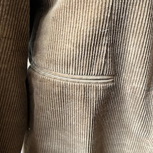 Op de afbeelding: Close-up van een bruine corduroy jas. De jas heeft een verticale geribbelde textuur en een zakdetail. De stof heeft een zacht, getextureerd uiterlijk. De jas lijkt een vintage of klassieke stijl te hebben.