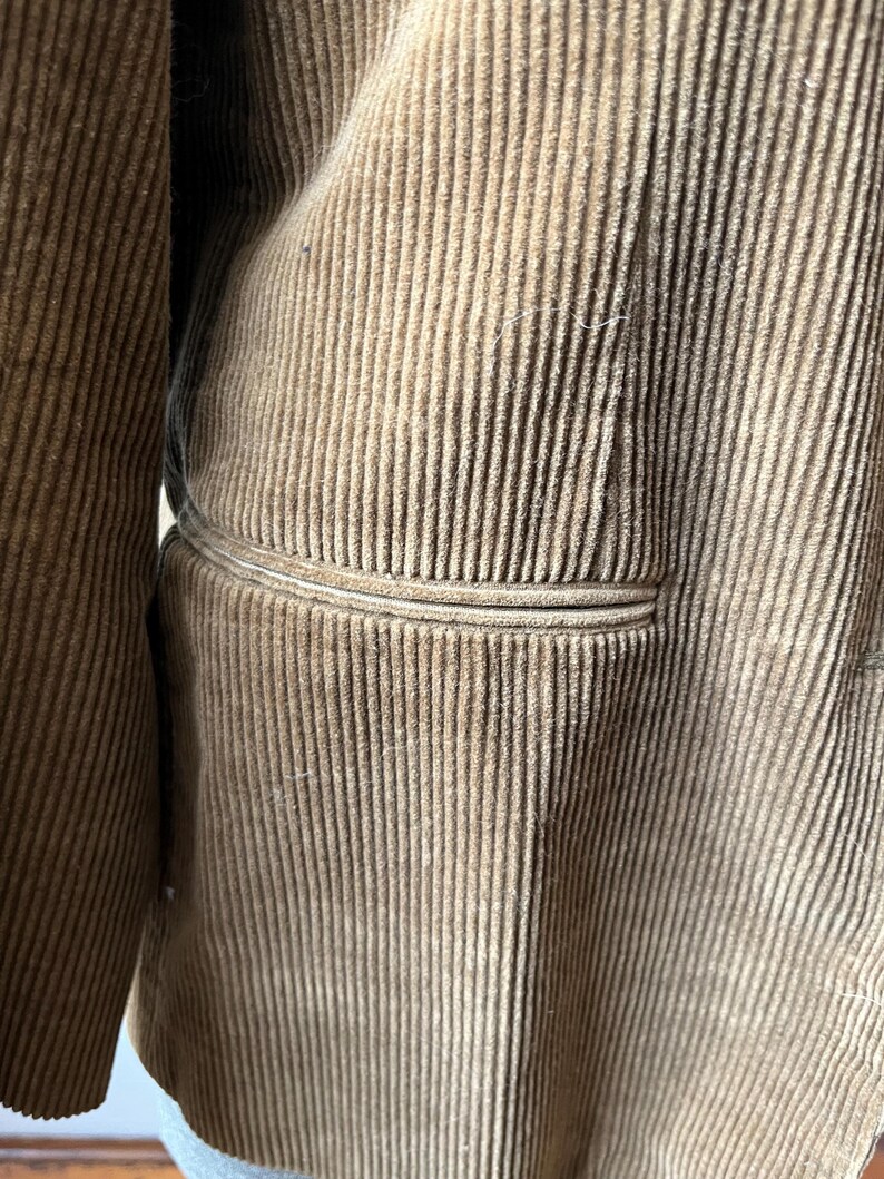 Op de afbeelding: Close-up van een bruine corduroy jas. De jas heeft een verticale geribbelde textuur en een zakdetail. De stof heeft een zacht, getextureerd uiterlijk. De jas lijkt een vintage of klassieke stijl te hebben.