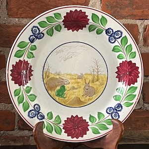 May include: A white ceramic plate with a hand-painted design of rabbits and a frog in a field. The plate has a green, blue, and red floral border.