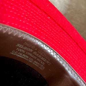 Puede incluir: Primer plano del interior de un sombrero de fieltro rojo con una banda de cuero marr&oacute;n. La banda tiene el texto "Filson Garment" y "100% Virgin Wool" impreso en ella.