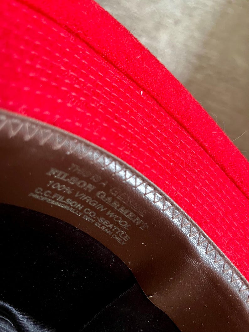 Puede incluir: Primer plano del interior de un sombrero de fieltro rojo con una banda de cuero marr&oacute;n. La banda tiene el texto "Filson Garment" y "100% Virgin Wool" impreso en ella.