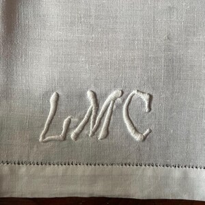 Puede incluir: Primer plano de un textil de lino blanco con las iniciales bordadas "LMC" en cursiva. La tela tiene una textura sutil y un dobladillo decorativo. El bordado está hecho con hilo blanco a juego.