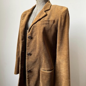 May include: A brown corduroy blazer with a notched lapel, three-button closure, and a chest pocket. The jacket is displayed on a mannequin and has two front pockets. The corduroy fabric has vertical ridges.