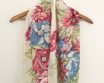 May include: A white silk scarf with a floral print featuring pink, blue, and green flowers. The scarf is folded and hanging on a wooden hanger.