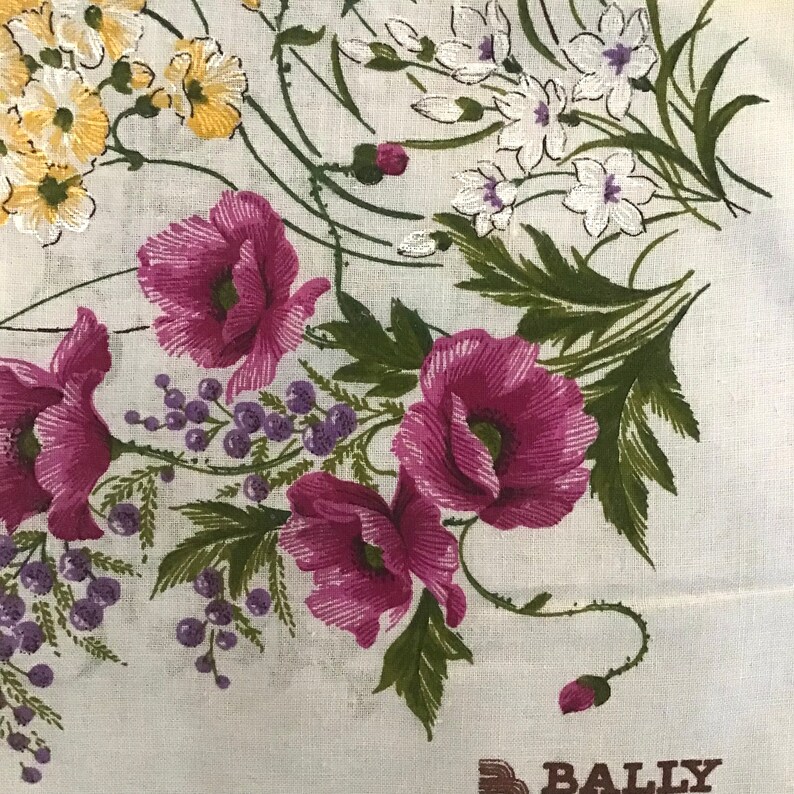 Pu&ograve; includere: Un tessuto bianco con un motivo floreale che presenta papaveri rosa, fiori bianchi e foglie verdi. Il tessuto ha la scritta "BALLY" stampata in lettere marroni in basso.