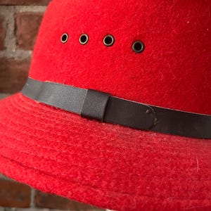 Puede incluir: Un sombrero de fieltro rojo con una banda de cuero marr&oacute;n y cinco ojales de metal. El sombrero tiene un ala ancha y una corona redonda.
