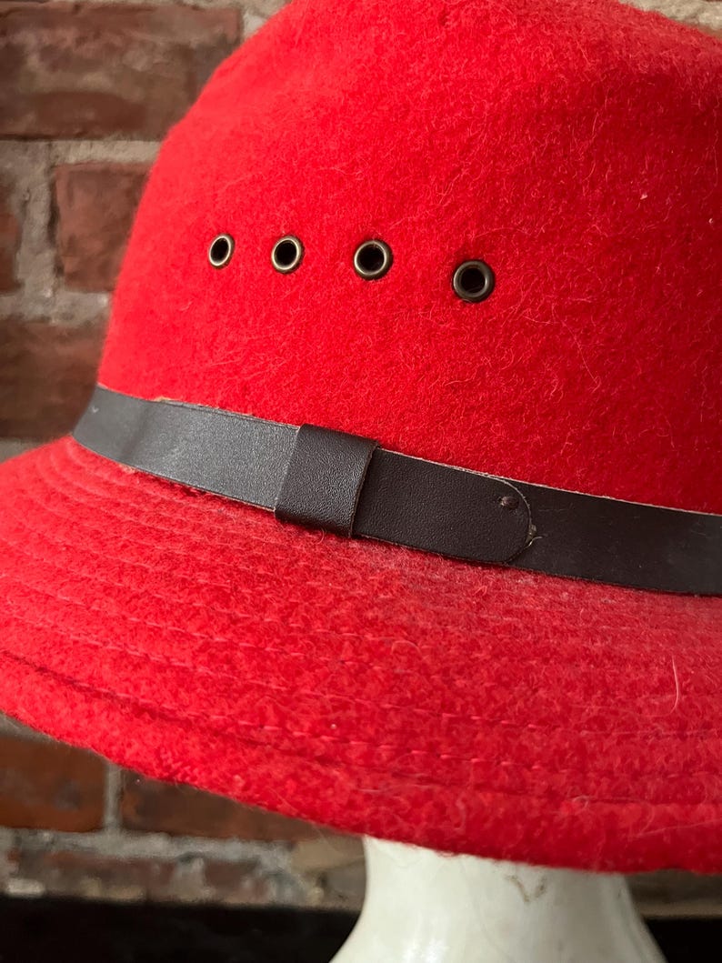 Puede incluir: Un sombrero de fieltro rojo con una banda de cuero marr&oacute;n y cinco ojales de metal. El sombrero tiene un ala ancha y una corona redonda.