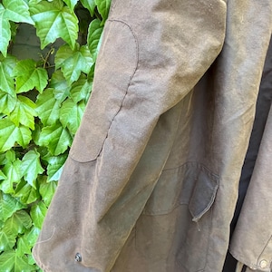 May include: Close-up of a brown waxed cotton coat. The coat features a patch on the elbow and a pocket. The coat is a dark brown color and has a button closure on the sleeve.