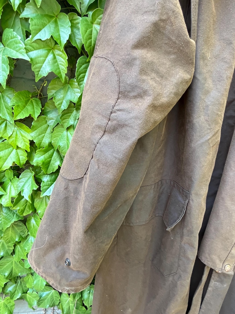 May include: Close-up of a brown waxed cotton coat. The coat features a patch on the elbow and a pocket. The coat is a dark brown color and has a button closure on the sleeve.