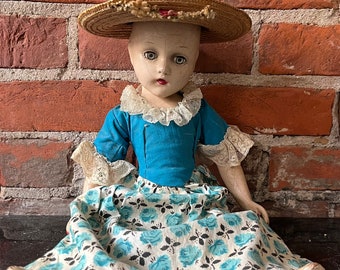 May include: A vintage porcelain doll wearing a blue dress with a floral print skirt and a straw hat. The doll has painted blue eyes and a painted smile.