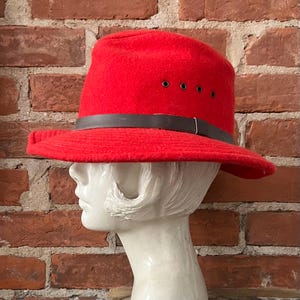 Puede incluir: Un sombrero fedora de fieltro rojo con una banda de cuero marr&oacute;n y cuatro ojales. El sombrero est&aacute; en una cabeza de maniqu&iacute; blanca.
