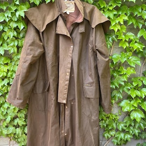 May include: A brown, full-length duster coat with a wide collar and button closures. The coat has two front pockets and a back vent. The coat is hanging on a white hanger against a backdrop of green ivy.