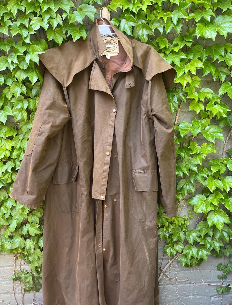 May include: A brown, full-length duster coat with a wide collar and button closures. The coat has two front pockets and a back vent. The coat is hanging on a white hanger against a backdrop of green ivy.