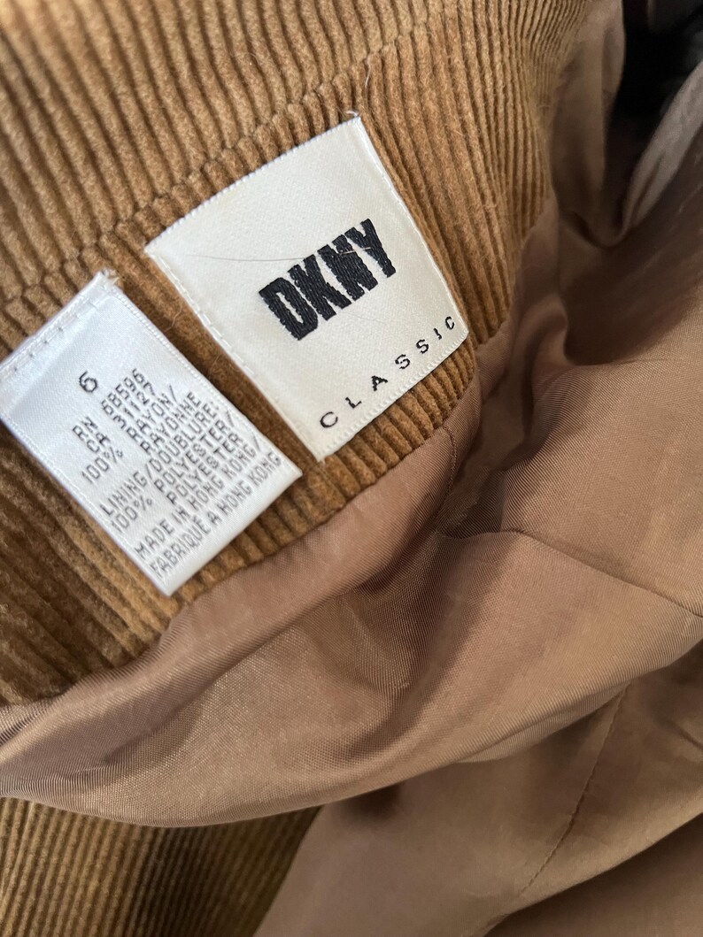 Op de afbeelding: Close-up van een bruin corduroy kledingstuk met een DKNY Classic label. Het label geeft aan dat het kledingstuk is gemaakt in Hong Kong en vermeldt de materialen rayon en polyester.