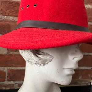 Puede incluir: Un sombrero fedora de fieltro rojo con una banda de cuero marr&oacute;n y ojales. El sombrero est&aacute; en una cabeza de maniqu&iacute; blanca.