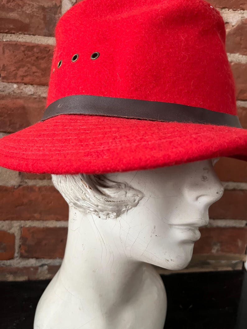 Puede incluir: Un sombrero fedora de fieltro rojo con una banda de cuero marr&oacute;n y ojales. El sombrero est&aacute; en una cabeza de maniqu&iacute; blanca.