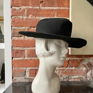 Peut inclure: Un chapeau fedora en feutre noir à large bord, posé sur une tête de mannequin blanche. Le chapeau est placé sur une surface noire avec un mur de briques en arrière-plan.