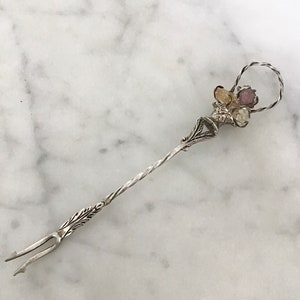 May include: A silver fork with a twisted handle and a decorative top with a cluster of colorful stones. The fork is designed for serving food.