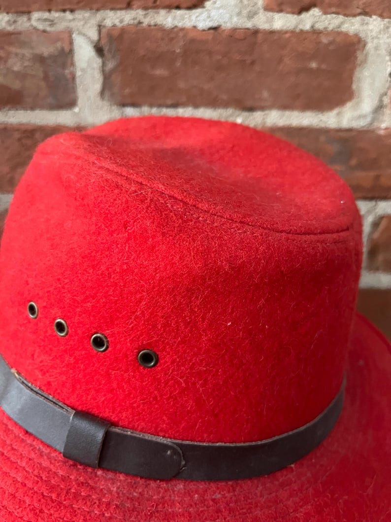 Puede incluir: Un sombrero fedora de fieltro rojo con una banda de cuero marr&oacute;n y cuatro ojales met&aacute;licos.