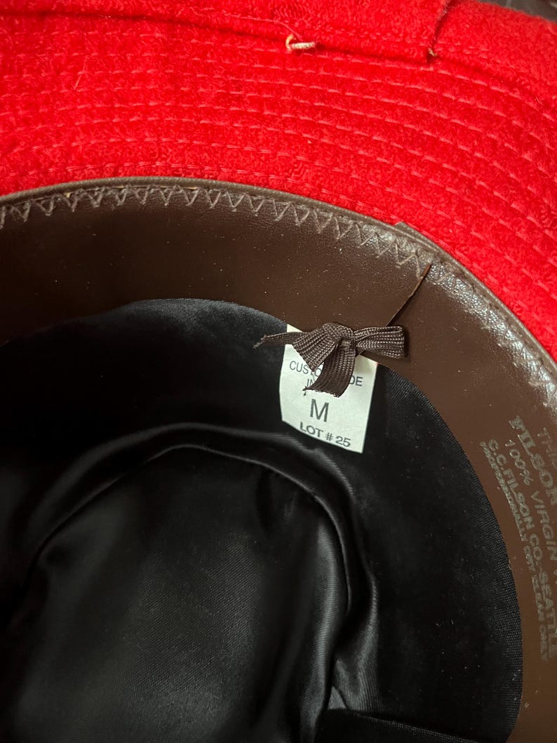Puede incluir: El interior de un sombrero de fieltro marr&oacute;n con una banda de fieltro rojo. El sombrero est&aacute; etiquetado con "FILSON 100% VIRGIN WOOL C.C. FILSON CO. SEATTLE" y tiene una etiqueta de talla que dice "M LOT #25".