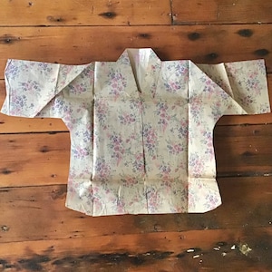 May include: A white blouse with a floral print in shades of pink and purple. The blouse has a V-neck and short sleeves.