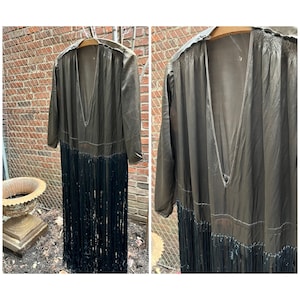 Può includere: Una lunga veste di colore marrone scuro, trasparente, con una profonda scollatura a V e un bordo di frange nere. La veste è appesa a un appendiabiti.