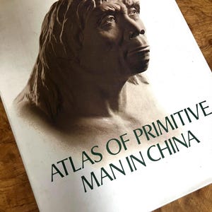 May include: A hardcover book titled Atlas of Primitive Man in China with a white cover featuring a sepia-toned illustration of a human bust with prominent facial features, looking upward. The title is printed in dark green capital letters.