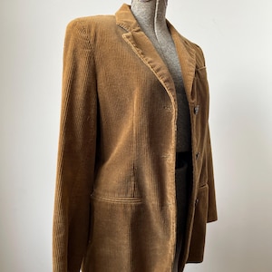 Op de afbeelding: Een bruine corduroy blazer tentoongesteld op een mannequin. De blazer heeft een revers, twee voorzakken en een sluiting met twee knopen. De stof heeft verticale ribbels en de kleur is een warme, aardse tint.