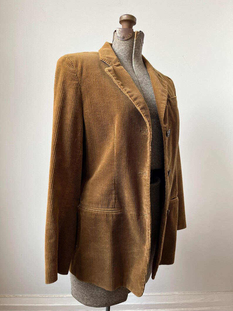 Op de afbeelding: Een bruine corduroy blazer tentoongesteld op een mannequin. De blazer heeft een revers, twee voorzakken en een sluiting met twee knopen. De stof heeft verticale ribbels en de kleur is een warme, aardse tint.
