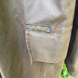 May include: Close-up of a brown garment with a pocket. A small rectangular label with the text "DRIZA-BONE" in gold lettering is sewn onto the pocket. The garment appears to be made of a durable, possibly waxed, fabric.