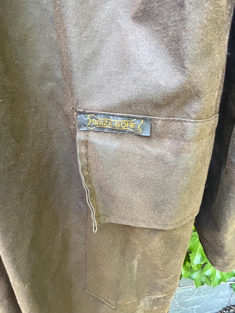 May include: Close-up of a brown garment with a pocket. A small rectangular label with the text "DRIZA-BONE" in gold lettering is sewn onto the pocket. The garment appears to be made of a durable, possibly waxed, fabric.