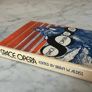 May include: A book titled "Space Opera" edited by Brian W. Aldiss. The cover has a blue and white illustration with orange and white stripes. The title is in large black letters.