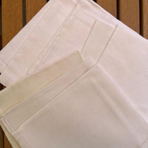 May include: A stack of off-white linen napkins with stitched edges. The napkins are folded and arranged on a wooden surface, showcasing their texture and design. The neutral color palette suggests a classic, elegant aesthetic, suitable for dining.